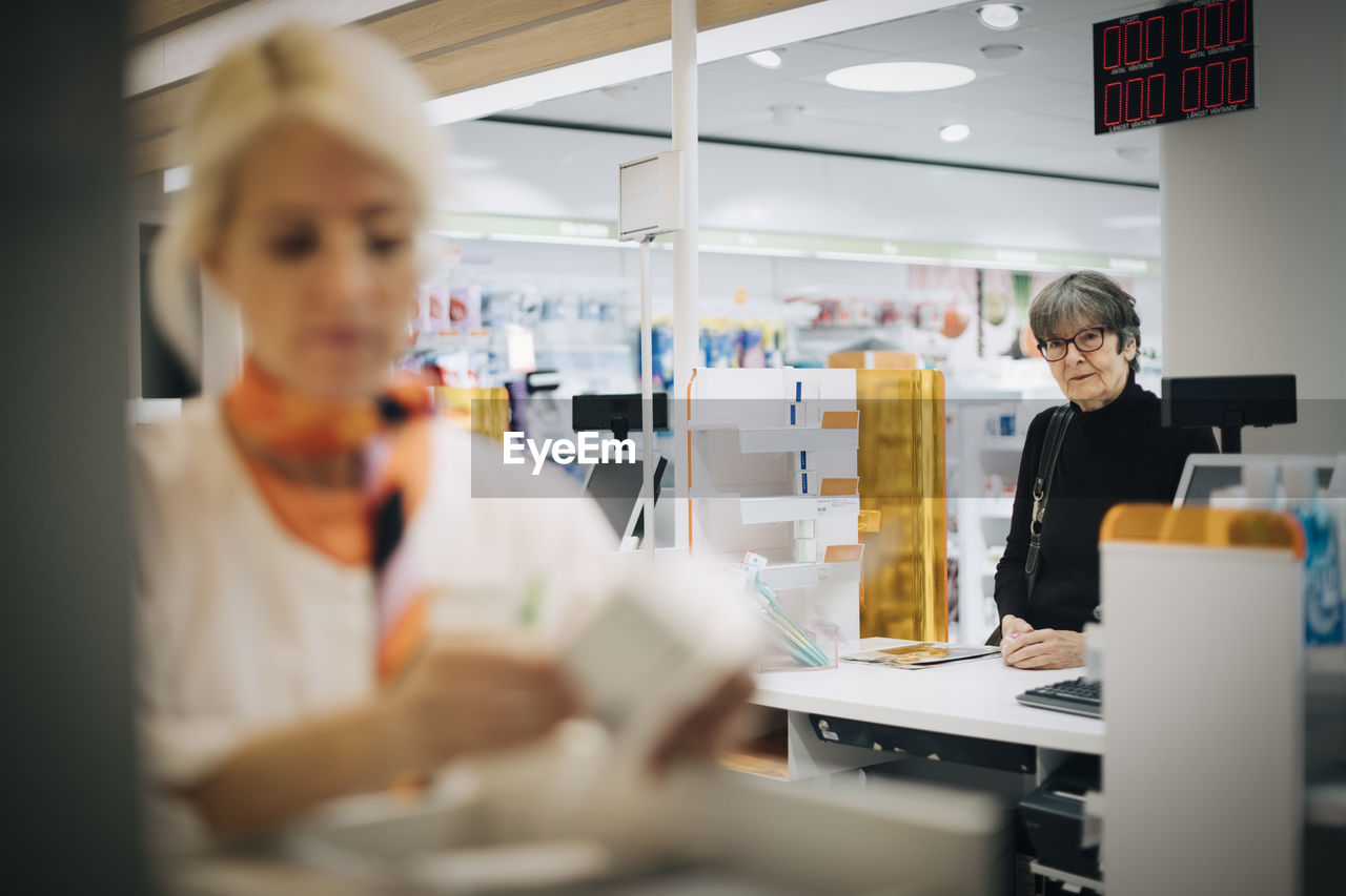 Senior female customer standing at checkout looking at owner in pharmacy store