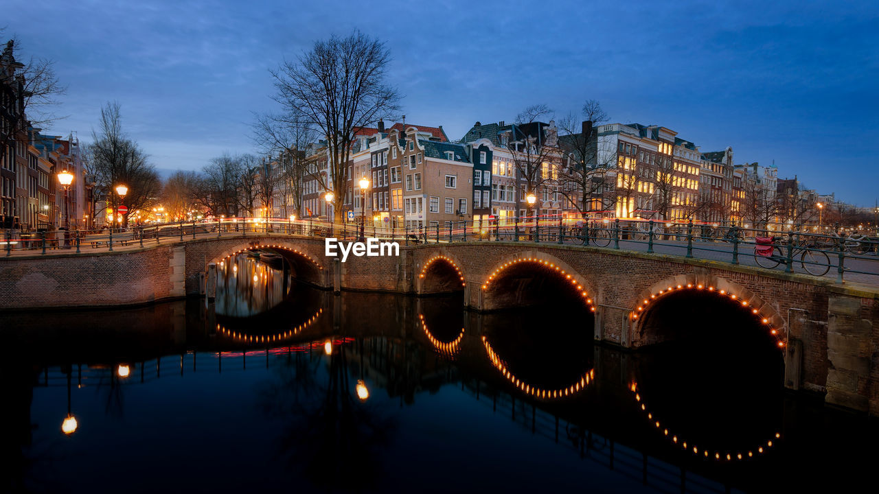 Illuminated bridge over river by buildings against sky at night