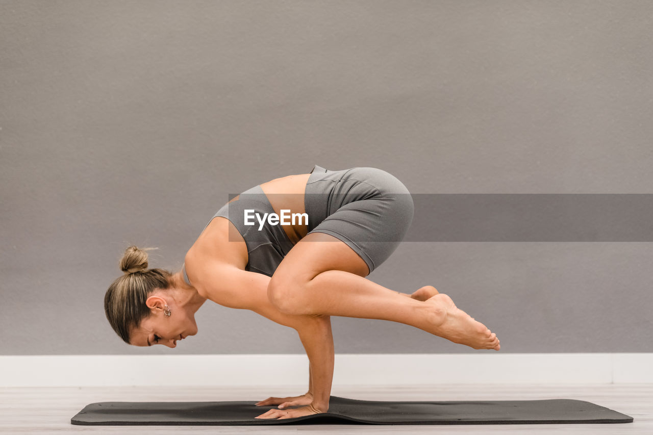 Side view of slim female in activewear balancing on hands in bakasana and practicing yoga on mat at home