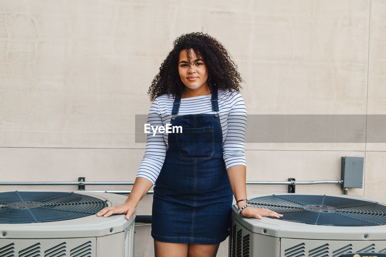 Portrait of young woman with curly hair standing against wall