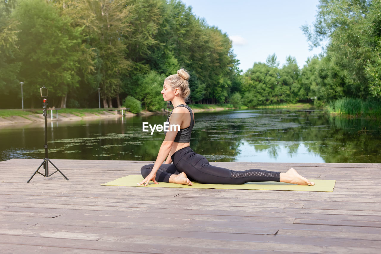 A beautiful woman in a on a wooden platform in summer, does yoga by pond in park