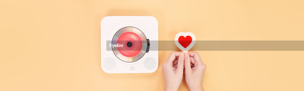 Hands holding heart shape red card next to white cd player on yellow 