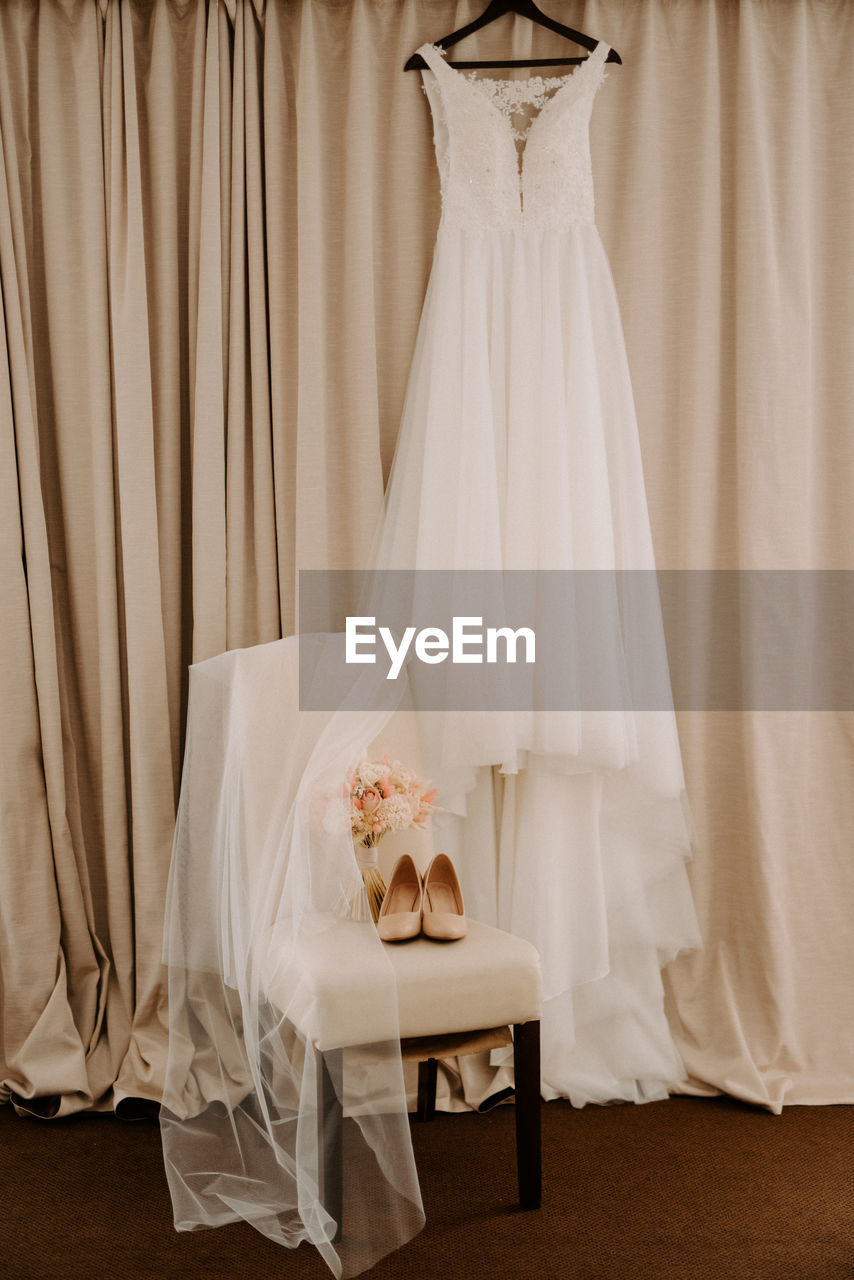 Wedding dress hanging on coat hanger against brown curtain