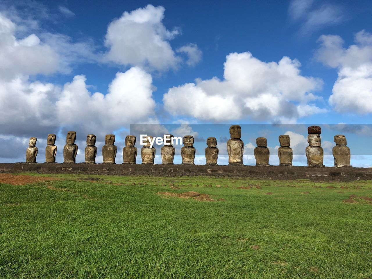 Low angle view of ancient moai statues by grassy land against cloudy sky