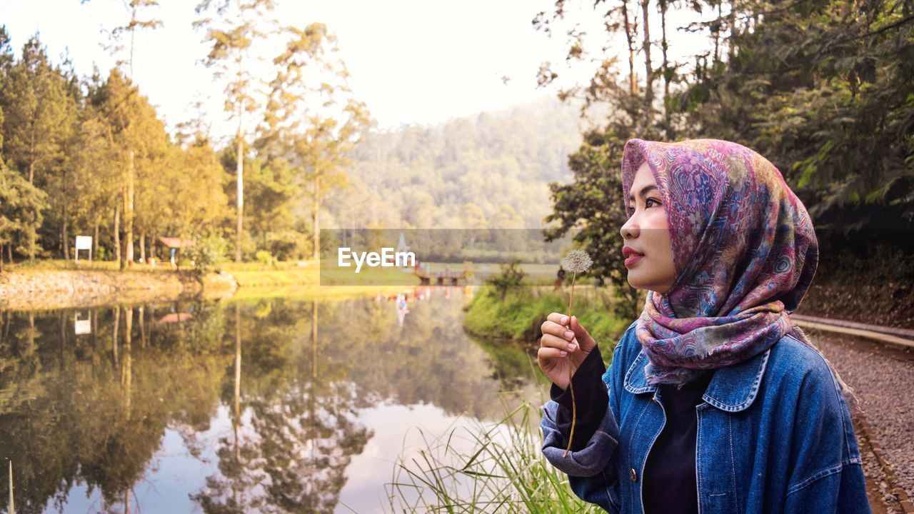 Side view of woman holding dandelion while standing by lake