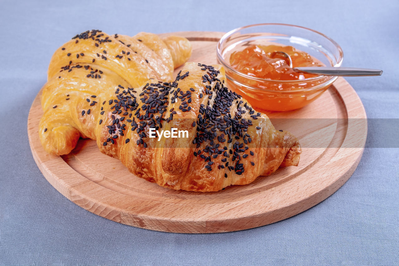 Two fresh croissants and glass bowl of jam are on  wooden cutting natural wood serving board 