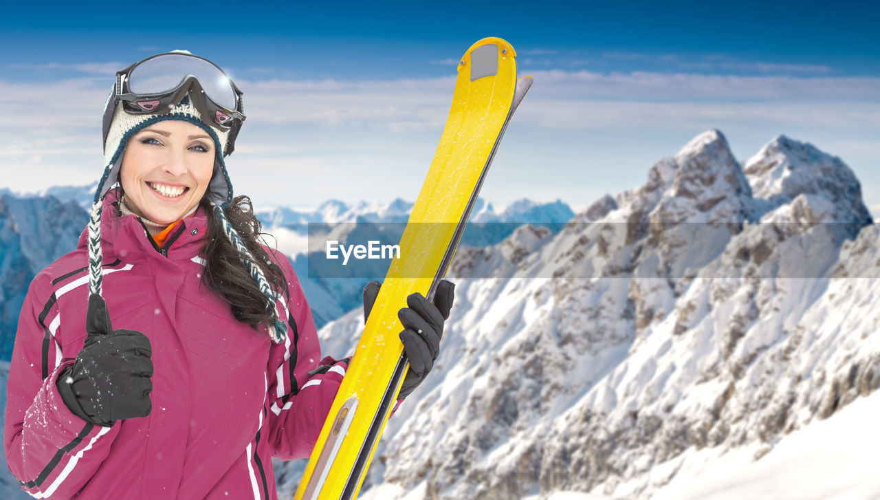 Portrait of cheerful woman holding skis while standing against snowcapped mountains