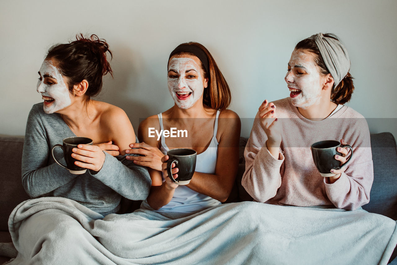 Cheerful female friends with facial masks holding mugs while sitting against wall at home