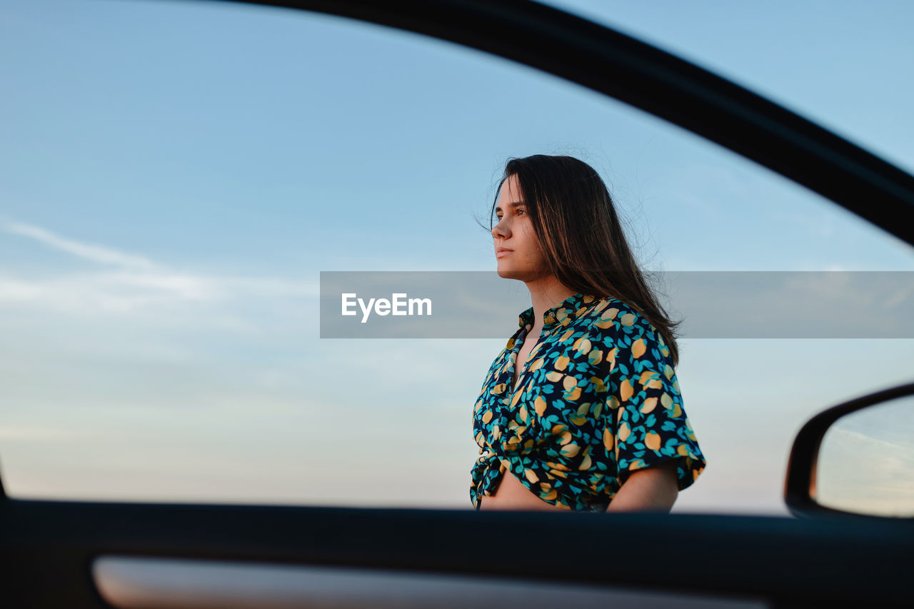 Young charming female in trendy wear with ornament looking away near auto door under blue cloudy sky