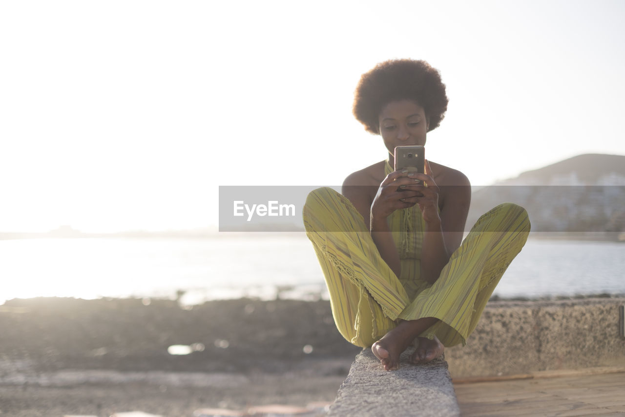 Young woman using mobile phone while sitting on retaining wall at beach against sky