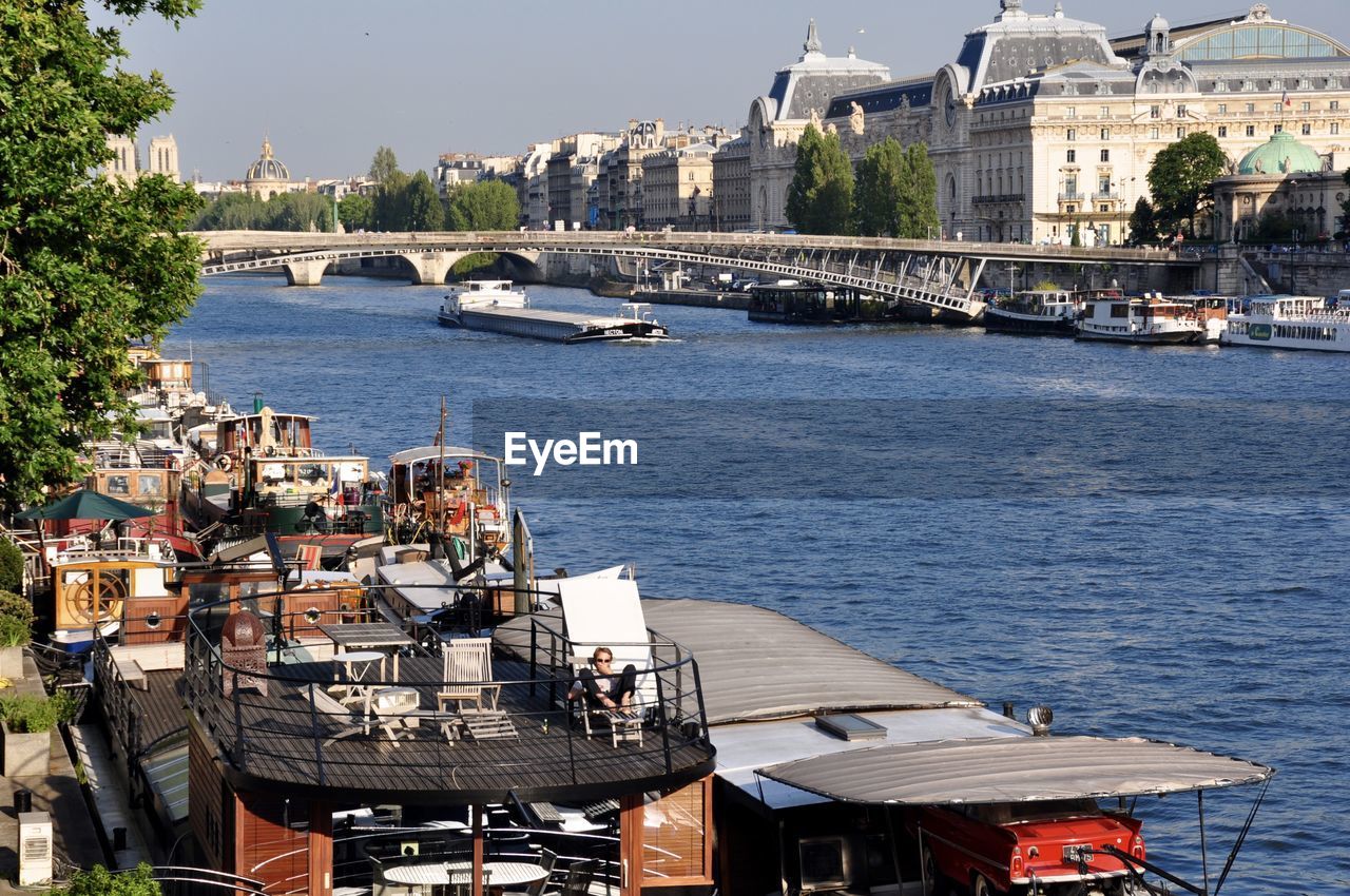 Boats moored in river by buildings on sunny day in city