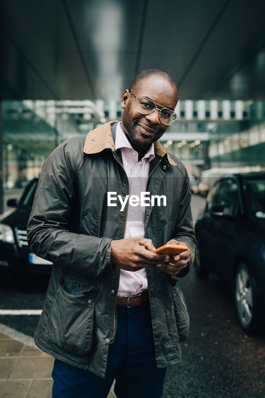 Portrait of businessman using smart phone while standing on road against building in city