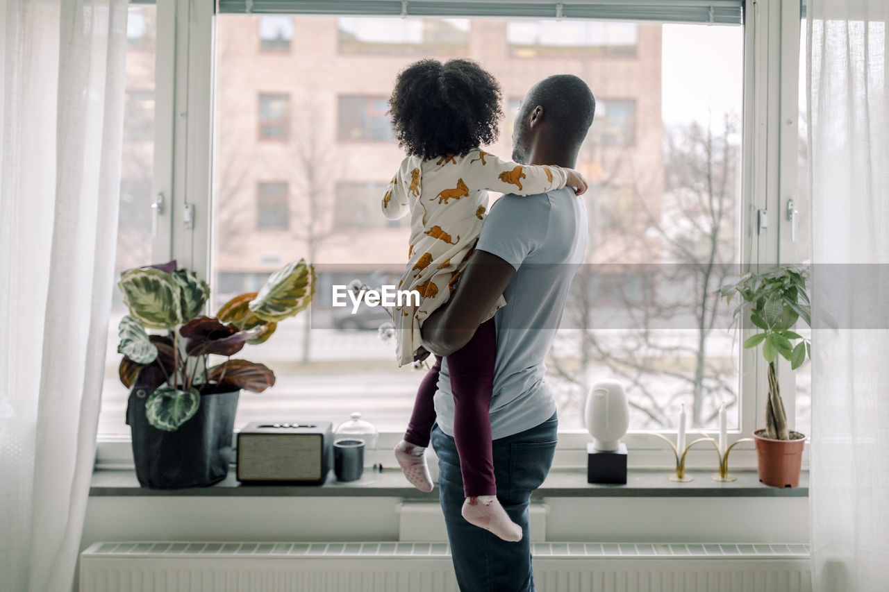 Father carrying daughter while looking through window at home
