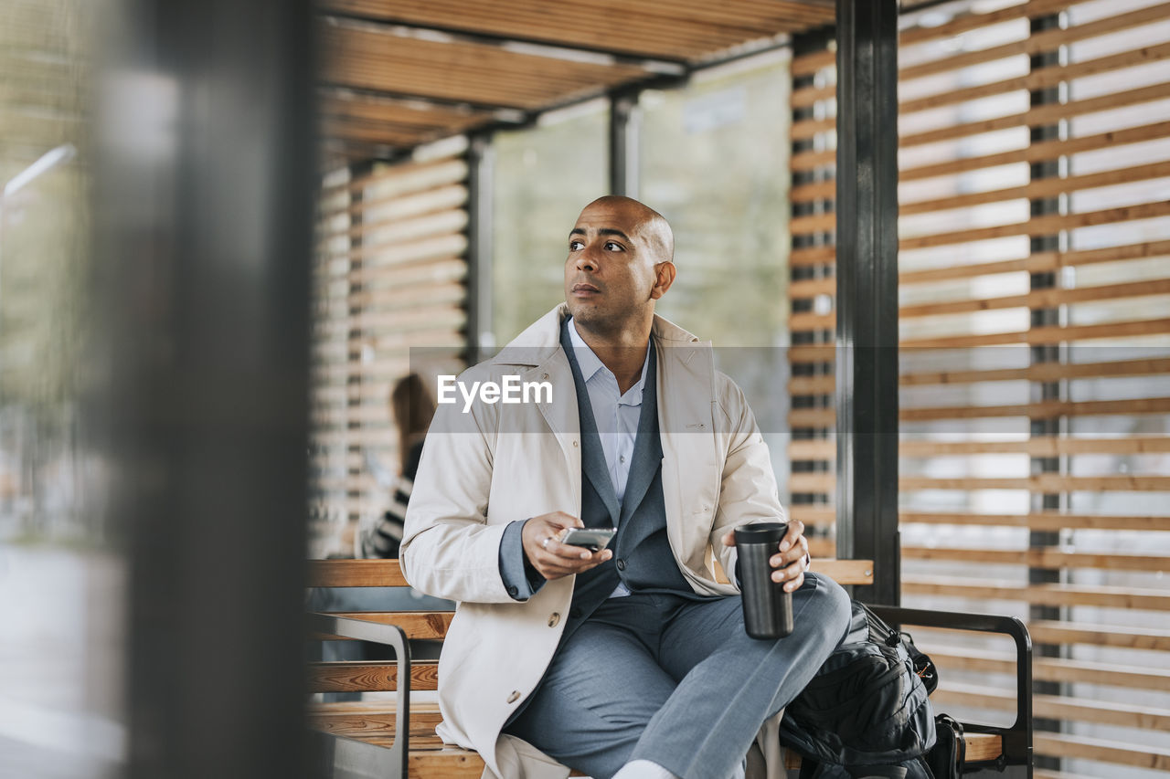 Businessman looking away holding smart phone and insulated drink container while sitting on bench at bus stop