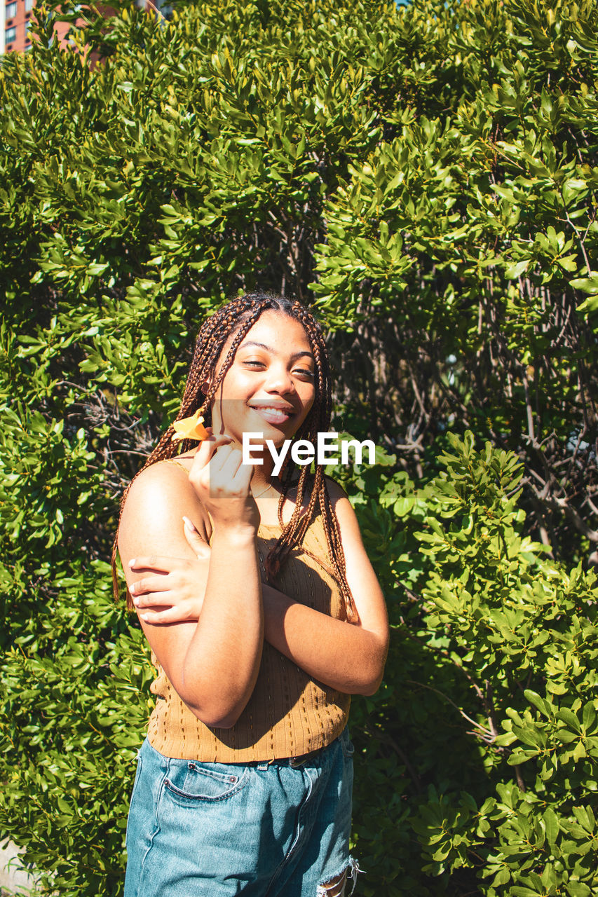 PORTRAIT OF A SMILING YOUNG WOMAN STANDING AGAINST PLANTS