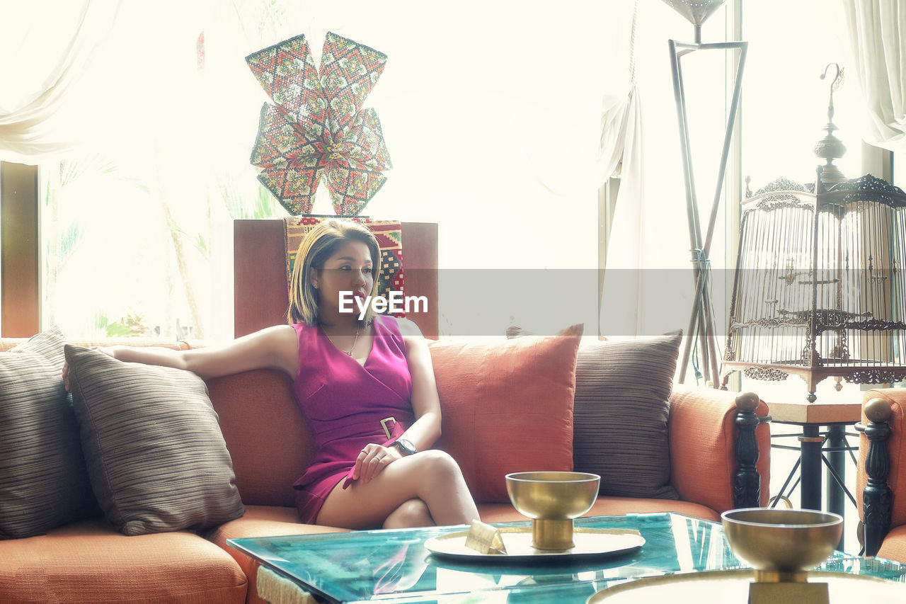 FULL LENGTH OF YOUNG WOMAN SITTING ON SOFA AT HOME