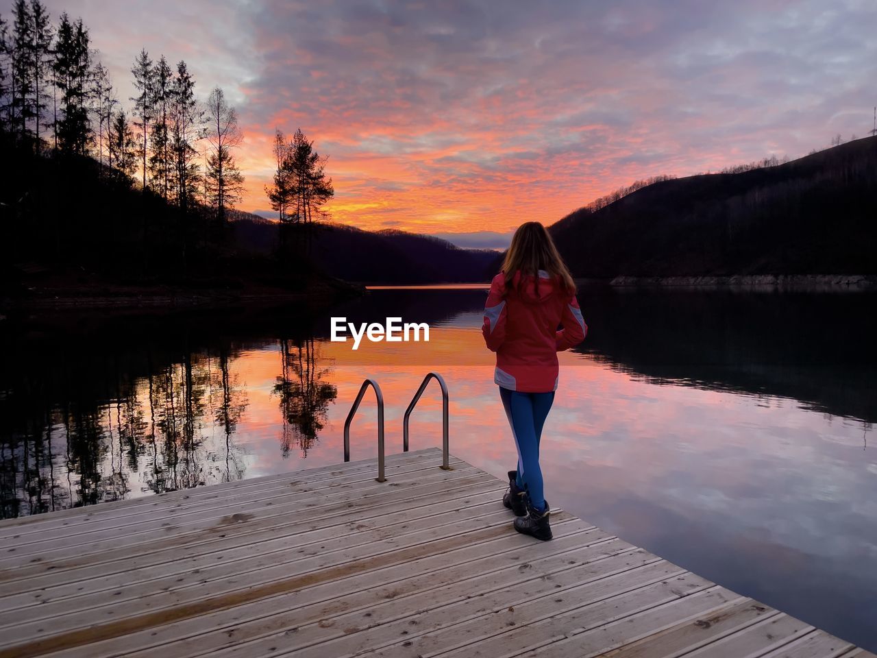 Woman standing on a wooden deck over the lake and watching an amazing sunset reflecting in the water