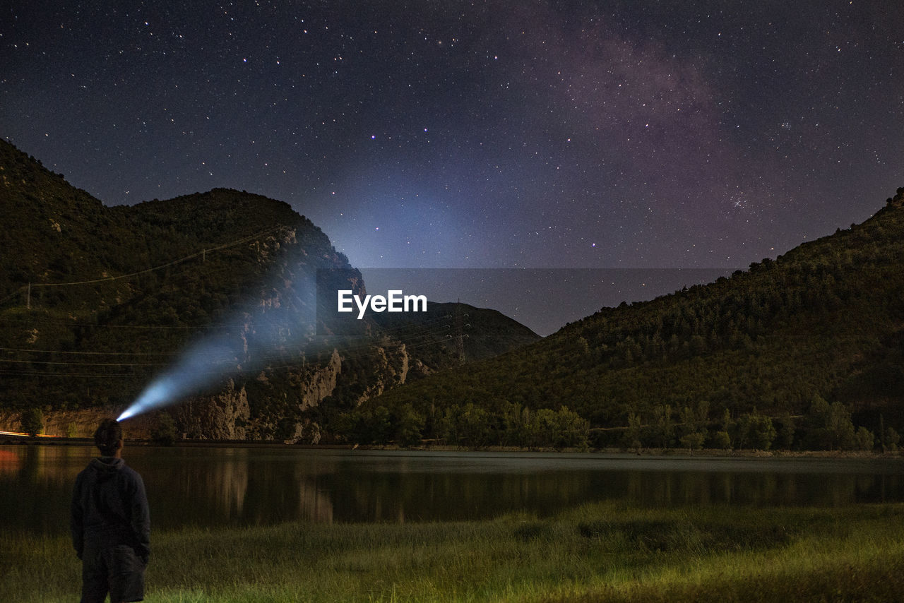 Rear view of man with illuminated headlamp on lakeshore against star field