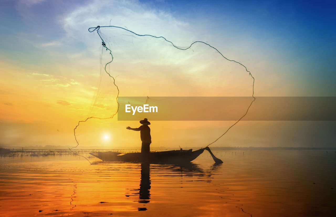 Silhouette fisherman standing on boat while throwing fishing net in sea against sky during sunrise