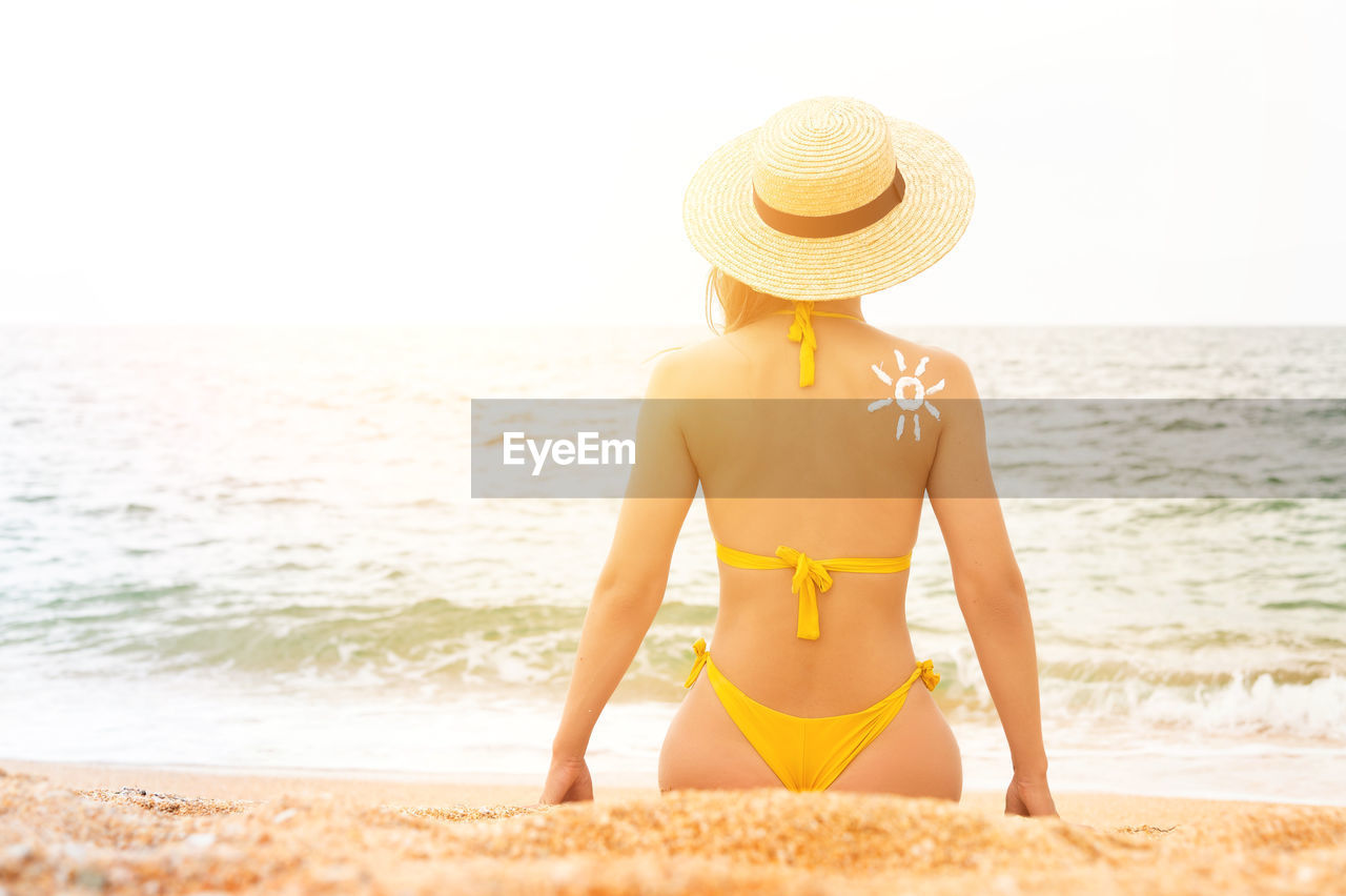 A young caucasian woman in a swimsuit and a straw hat sits on the beach in front of the sea with her