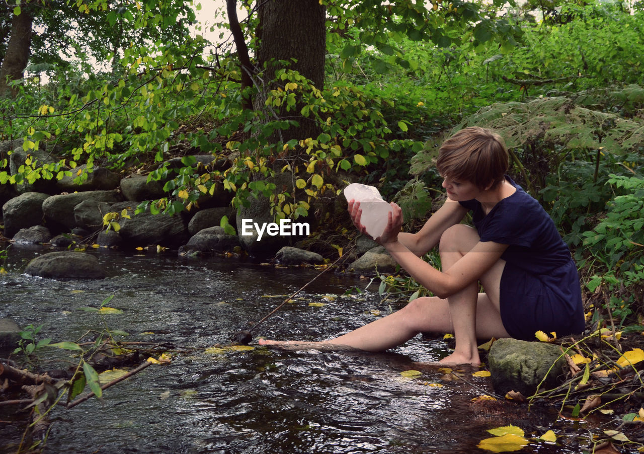 Young woman holding ice while sitting by stream in forest