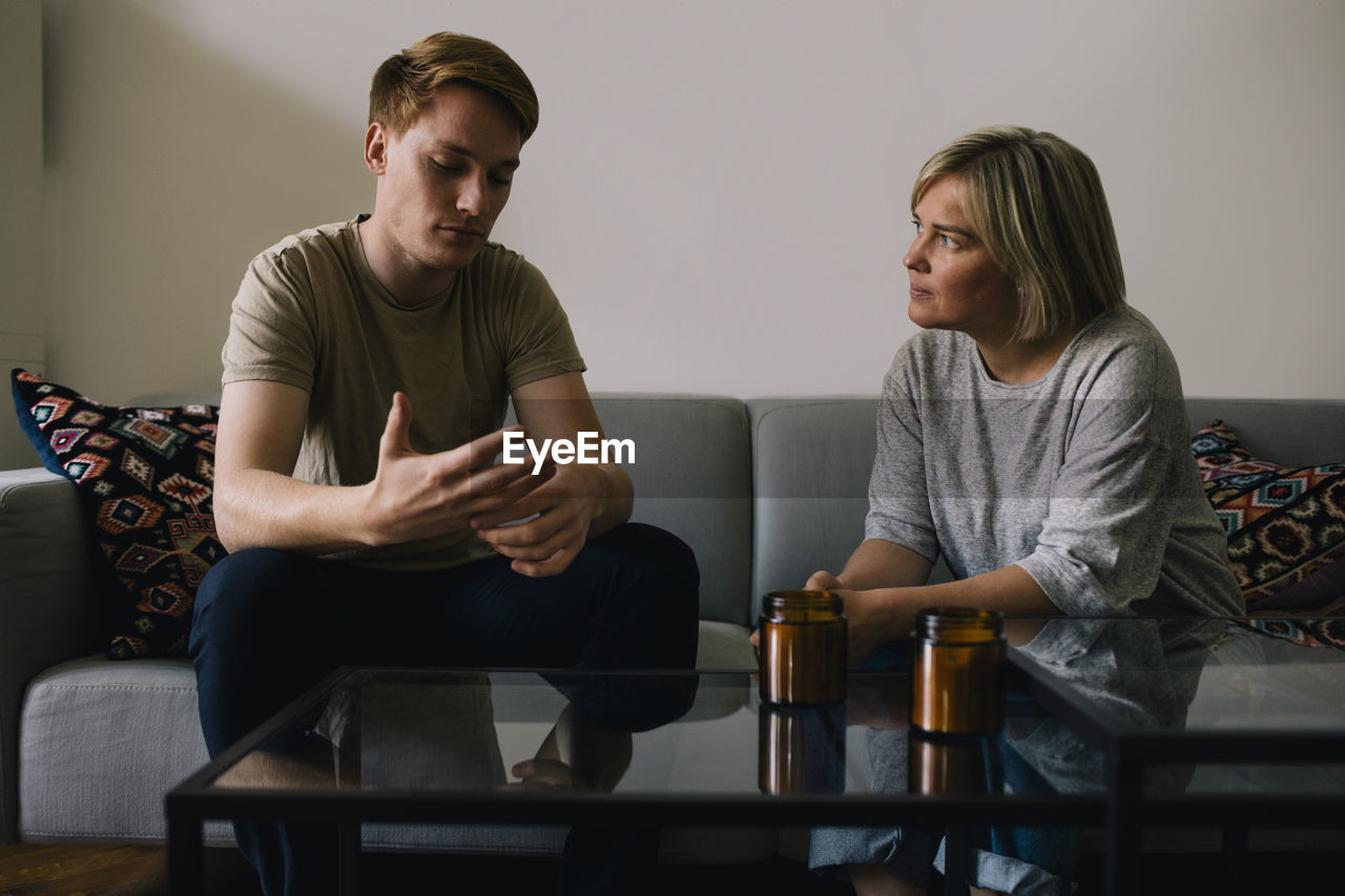 Mature parent looking at sad son sitting on sofa at home