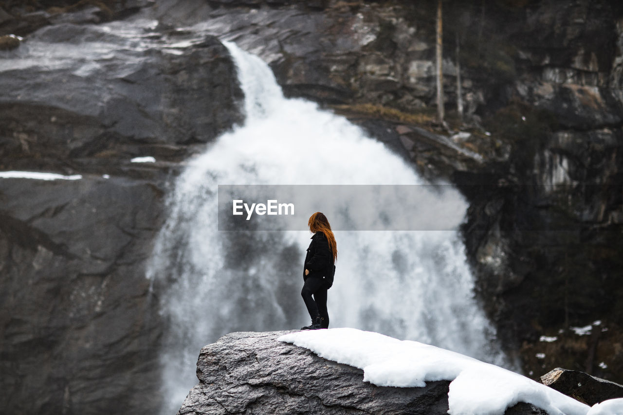 Side view of traveling woman standing on rock and observing amazing view of powerful waterfall in highlands in winter