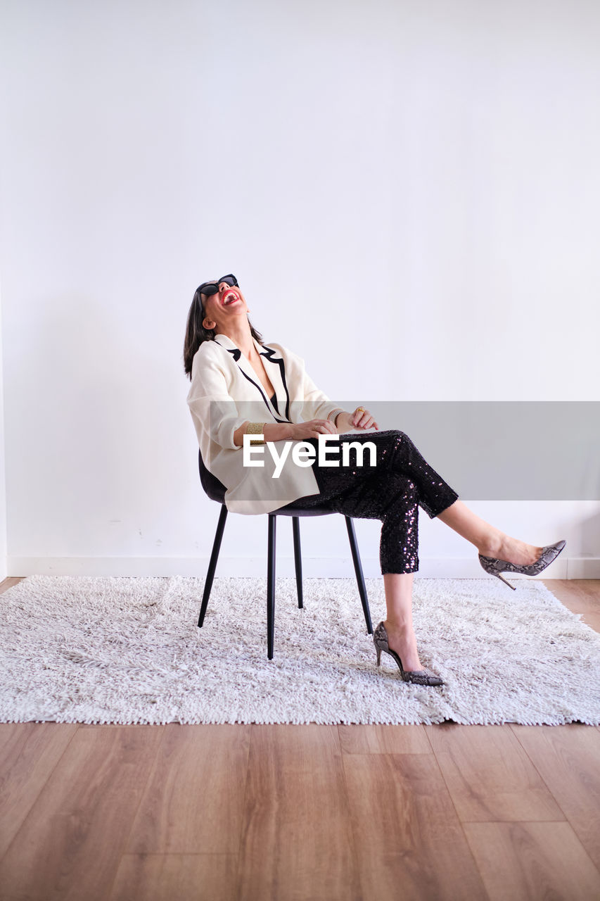 Full body of positive female in trendy outfit and high heels sitting on rug near white wall in light studio