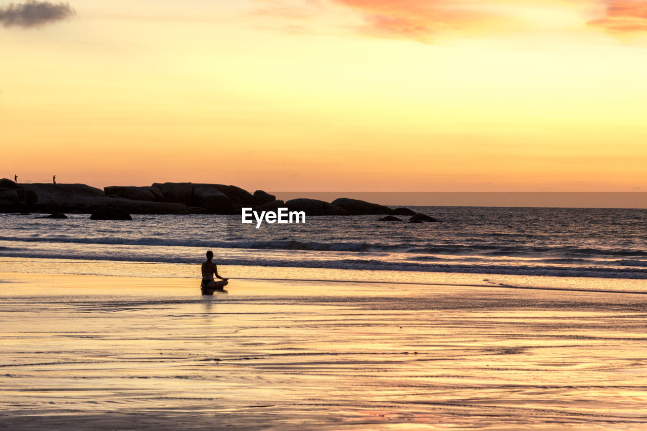 Silhouette person meditating at beach against sky during sunset