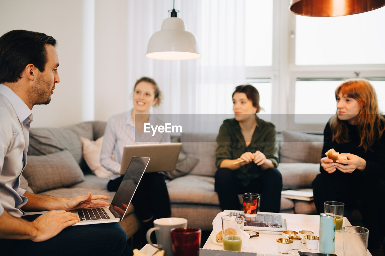 Female professionals looking at businessman sitting with laptop on sofa in creative office