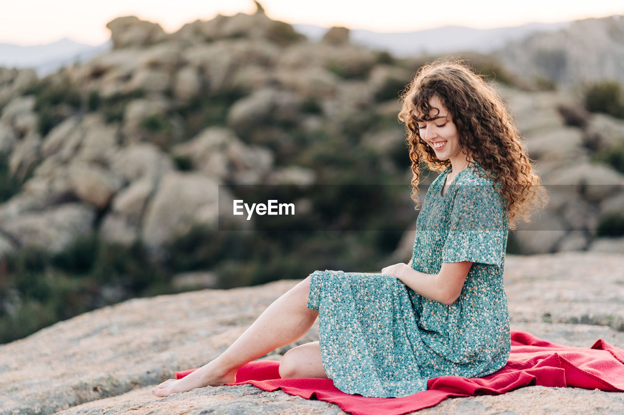 Young barefoot female in blue maxi sundress sitting on red plaid on rough rocky hilltop and looking down