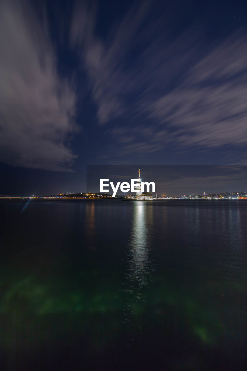 Illuminated city at waterfront against cloudy sky