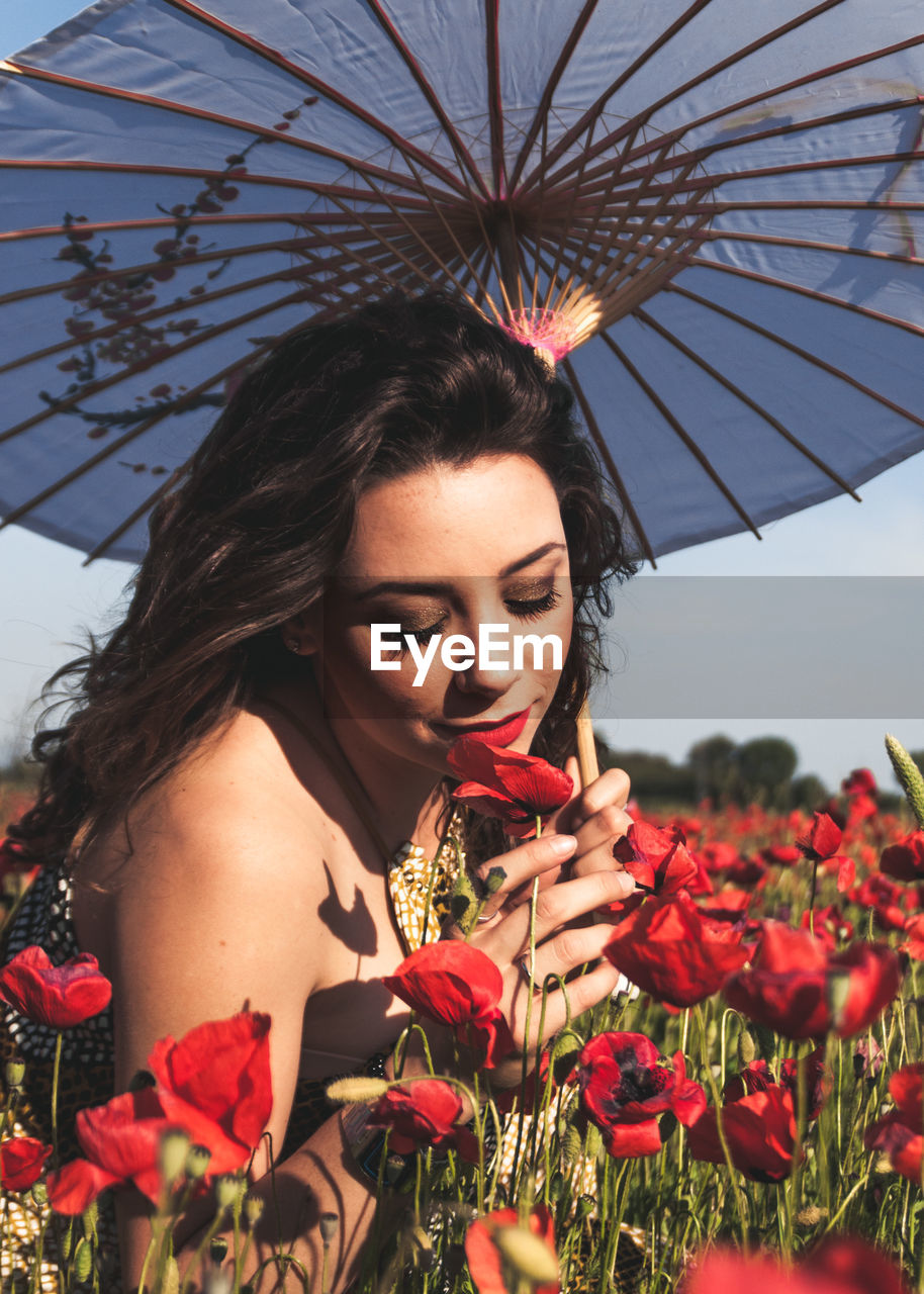 Young woman with umbrella smelling red flowers during sunny day