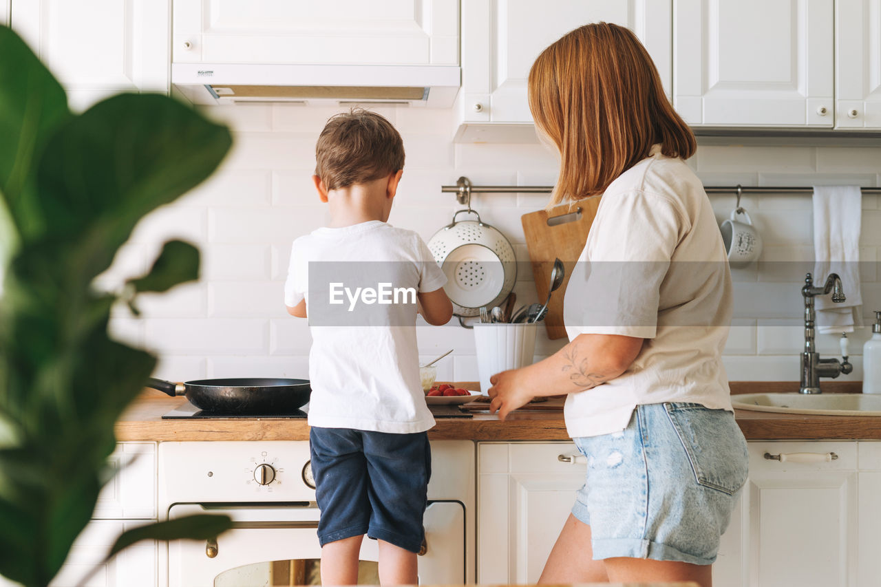 Cute toddler boy with mother cooking breakfast with puncakes together in bright kitchen at home