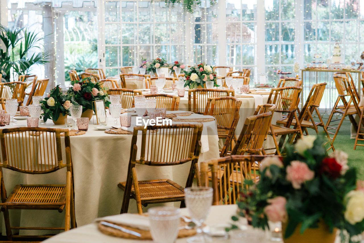 Big spacious room with festive decorated tables and wooden chairs under ceiling with green plants