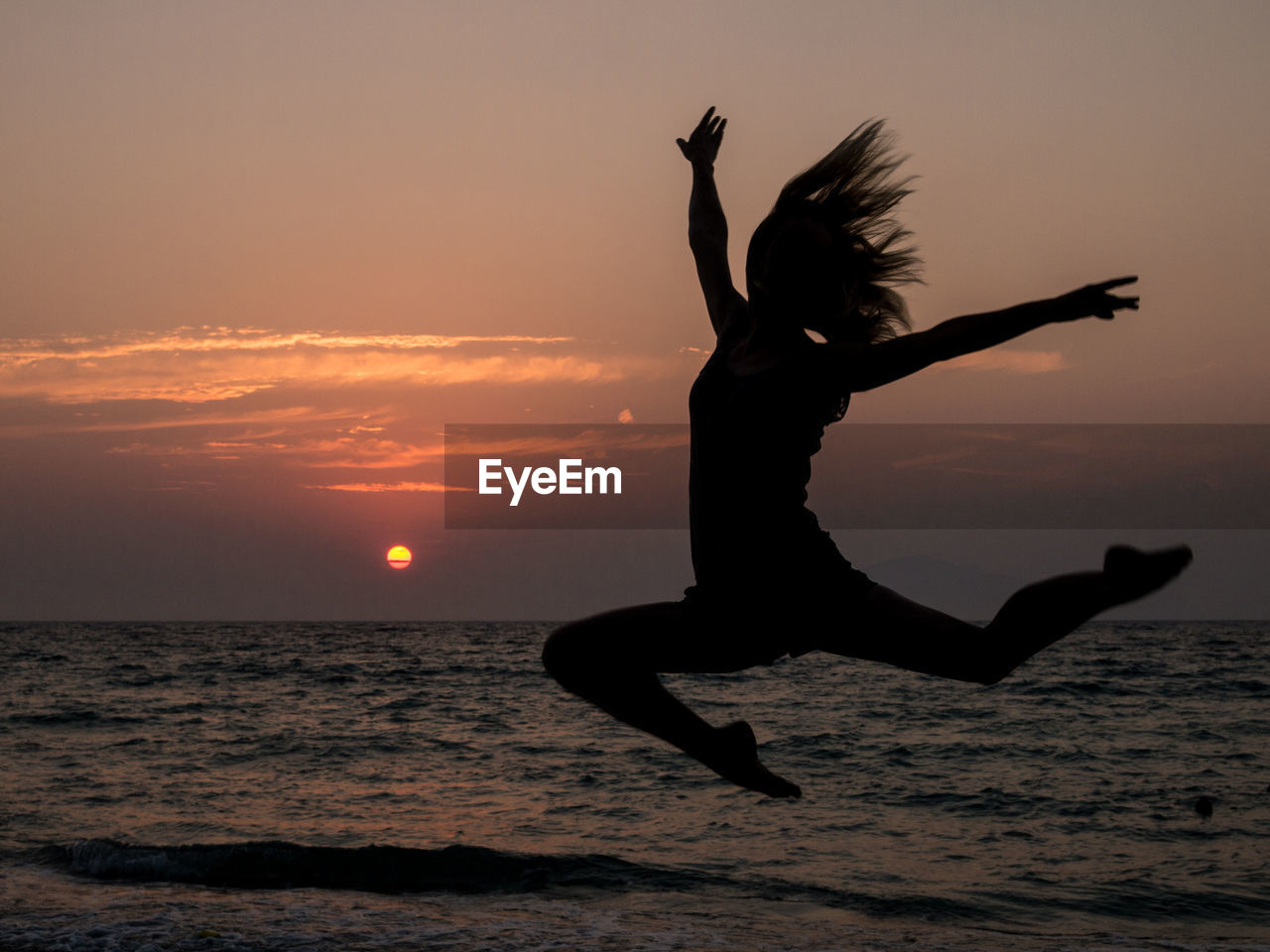 Silhouette woman with hand raised jumping at beach against sky during sunset