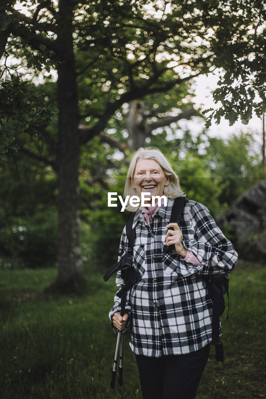 Portrait of happy senior woman holding hiking poles while hiking in forest