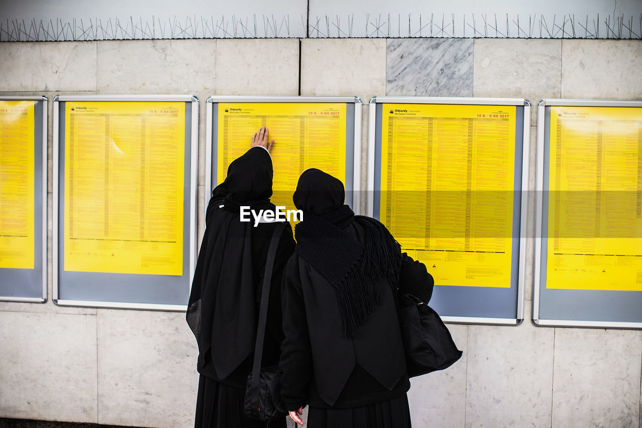 REAR VIEW OF TWO WOMEN STANDING AGAINST YELLOW WALL