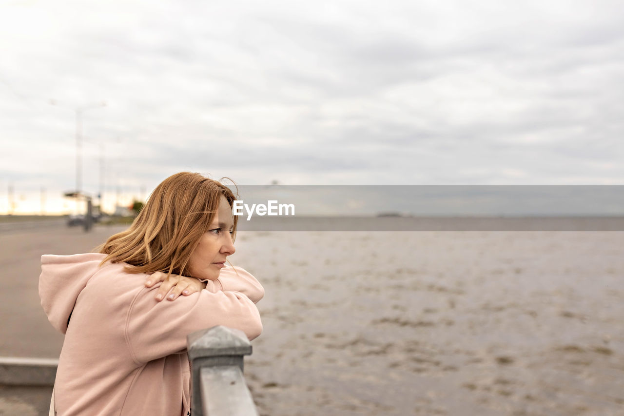 REAR VIEW OF WOMAN STANDING ON BEACH AGAINST SKY