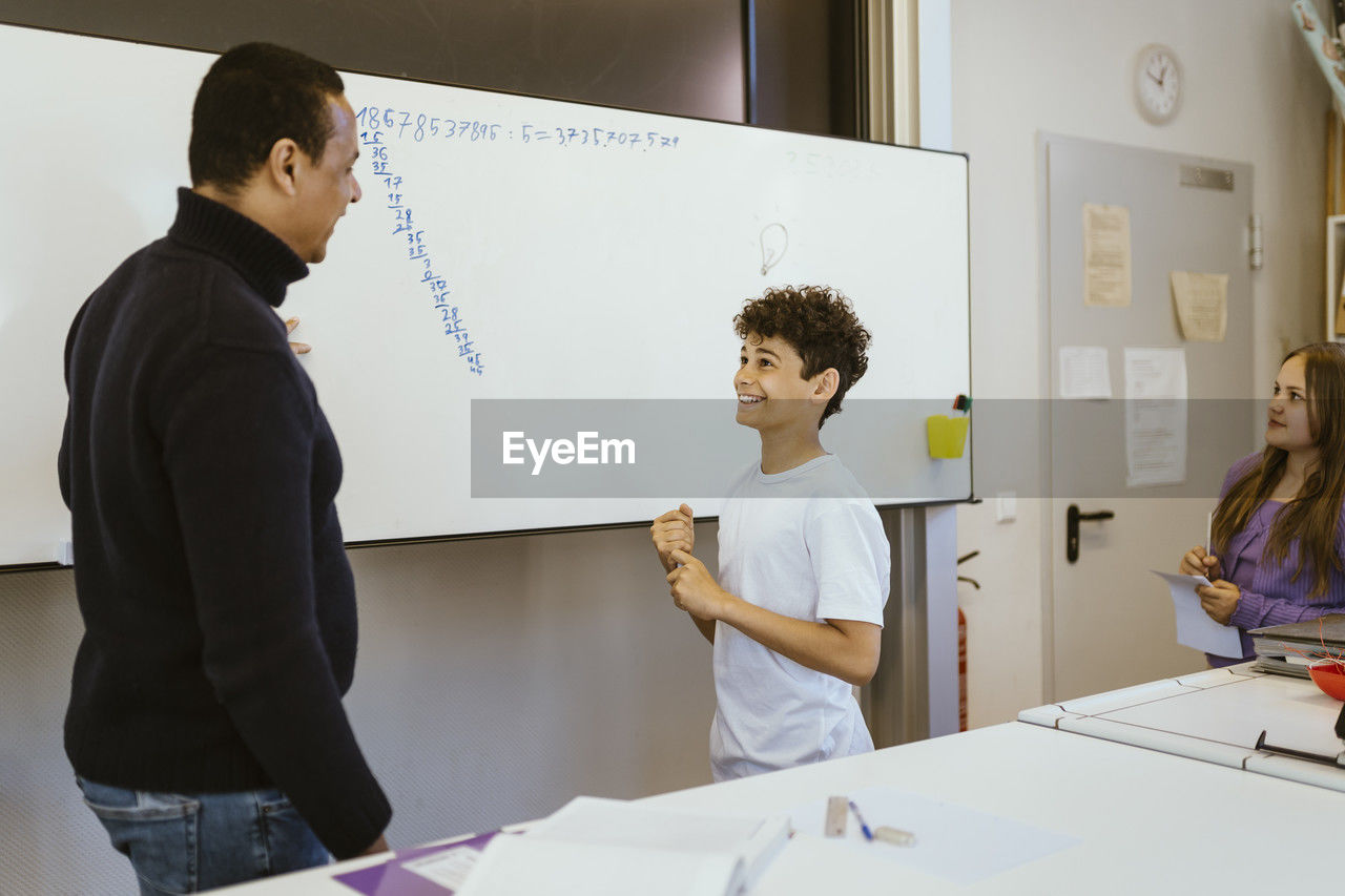 Smiling boy talking to male teacher standing near whiteboard in classroom