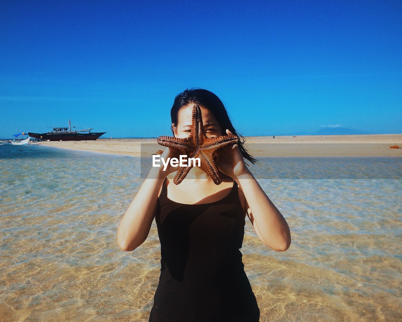 Woman covering face with dead starfish on shore against blue sky