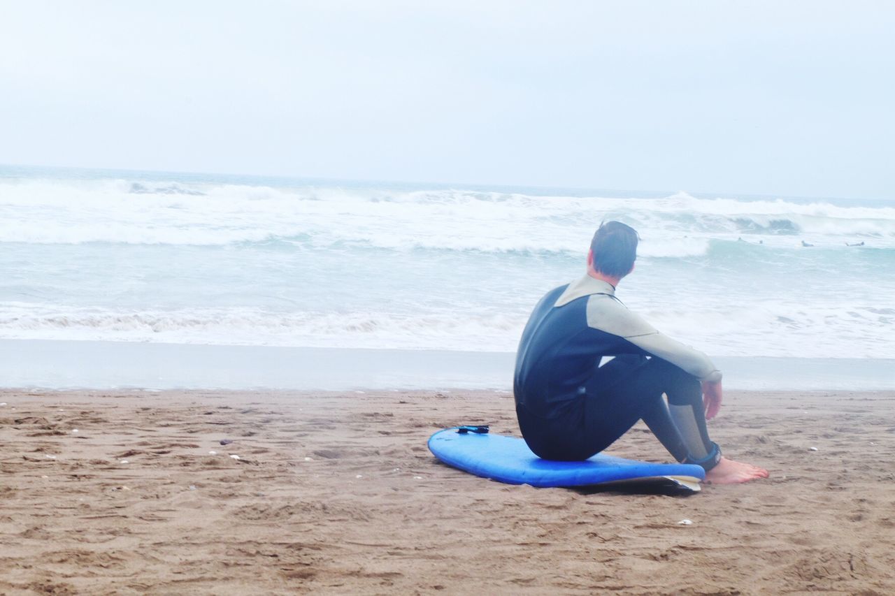 Man with surfboard sitting on shore at beach against sky