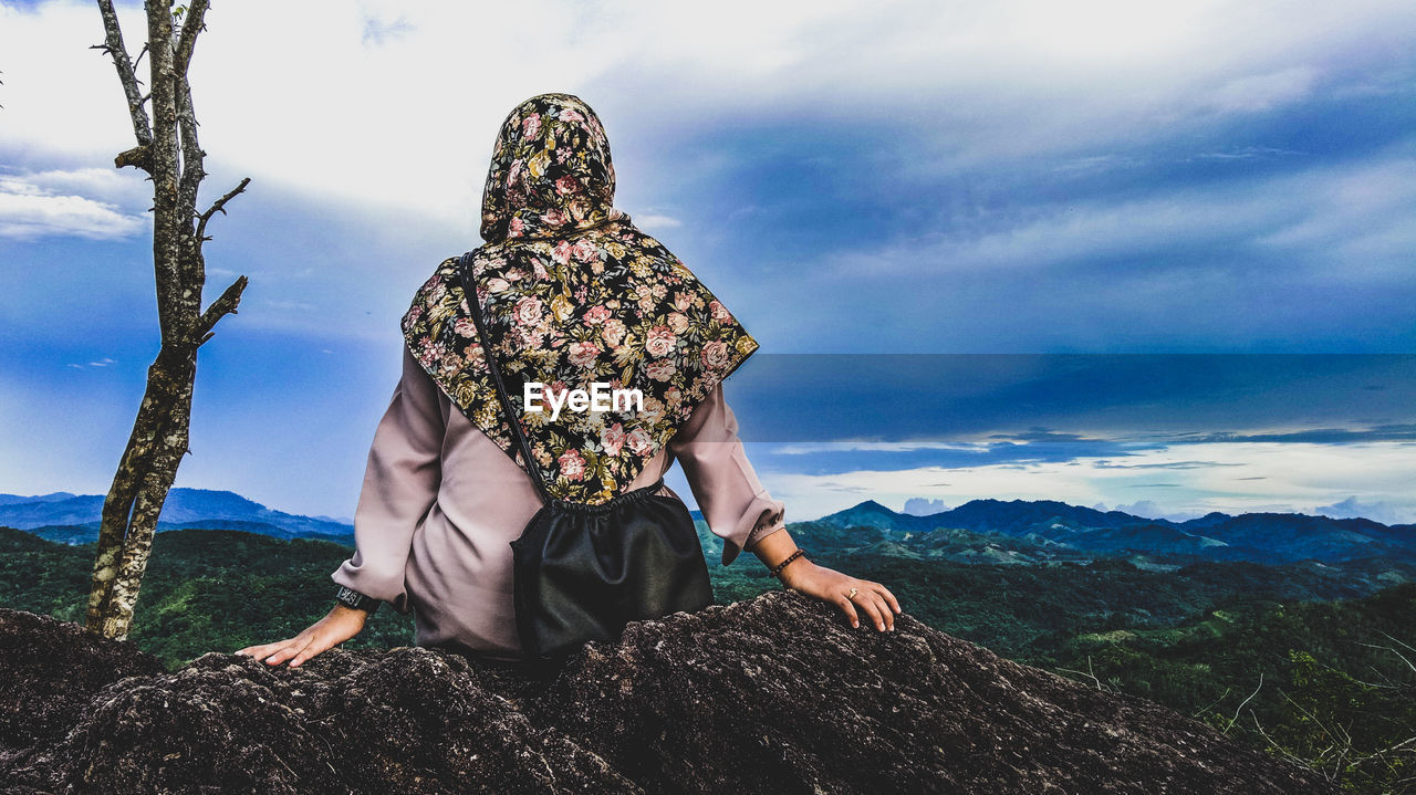 Rear view of woman sitting on cliff against cloudy sky