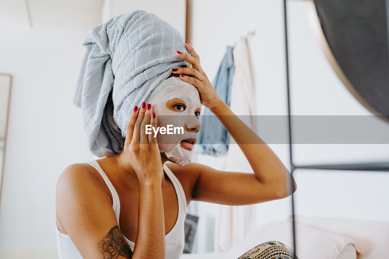 Young female in mirror applying skincare moisturizing cloth mask on face during home beauty procedure