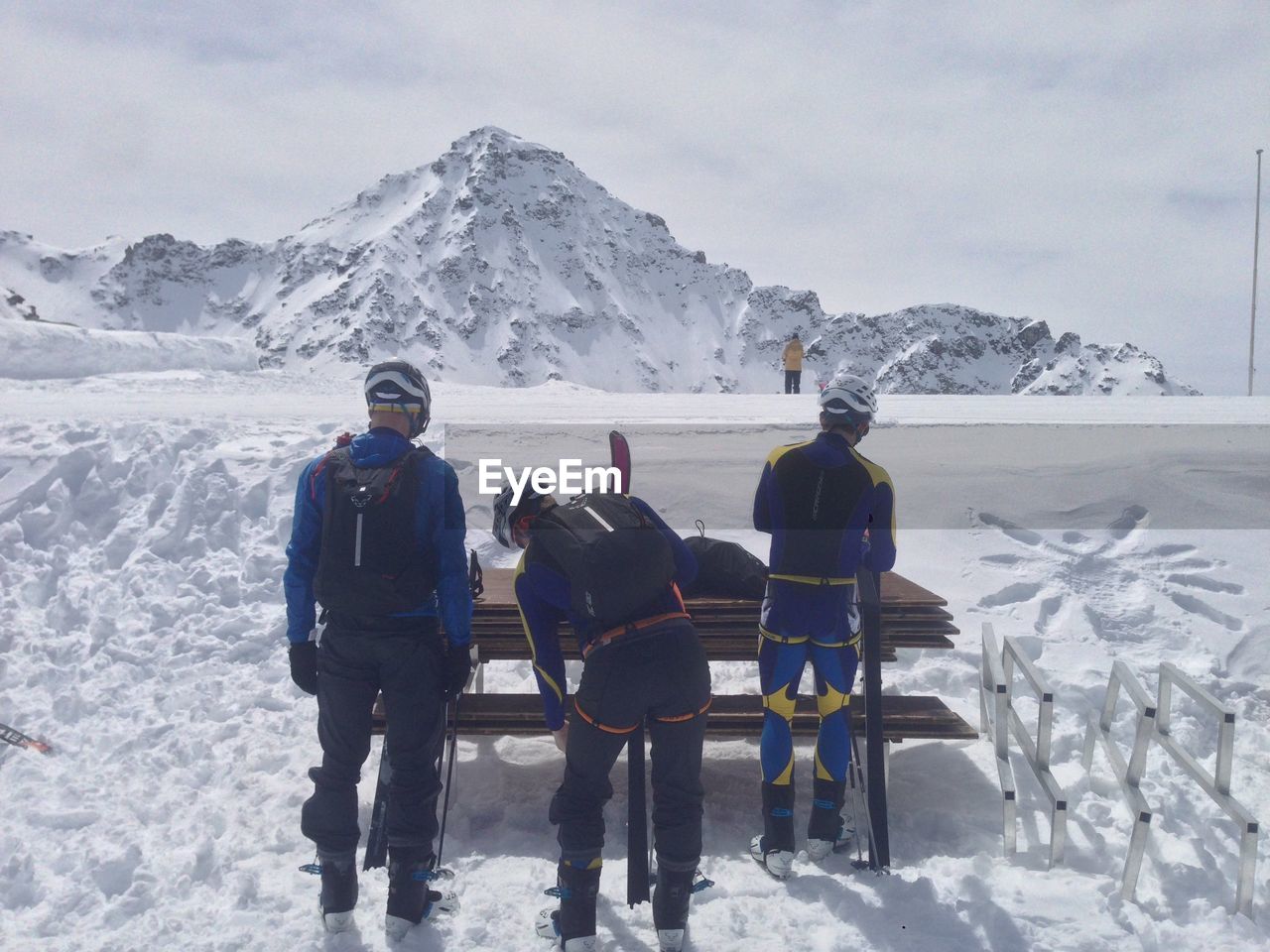 Rear view of skiers standing by wooden bench on snow covered field against mountain