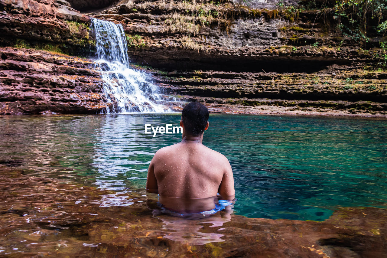 Young man sitting in natural waterfall clear water at morning from flat angle