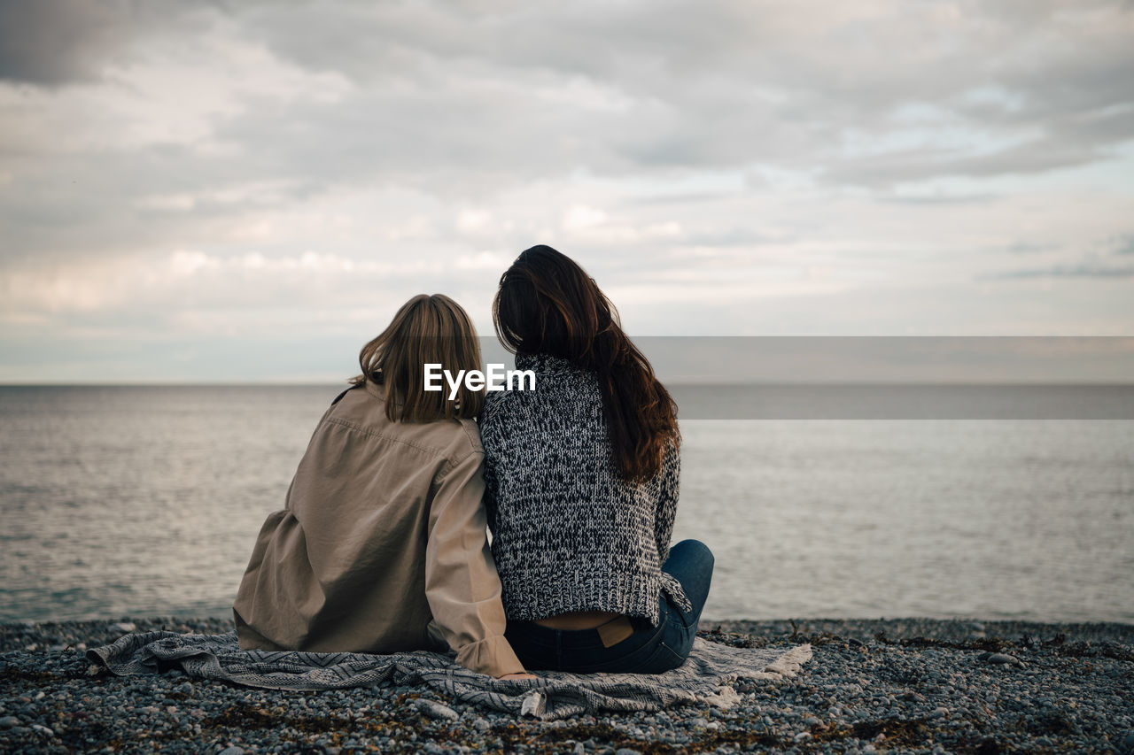 Rear view of female friends spending leisure time at beach against cloudy sky