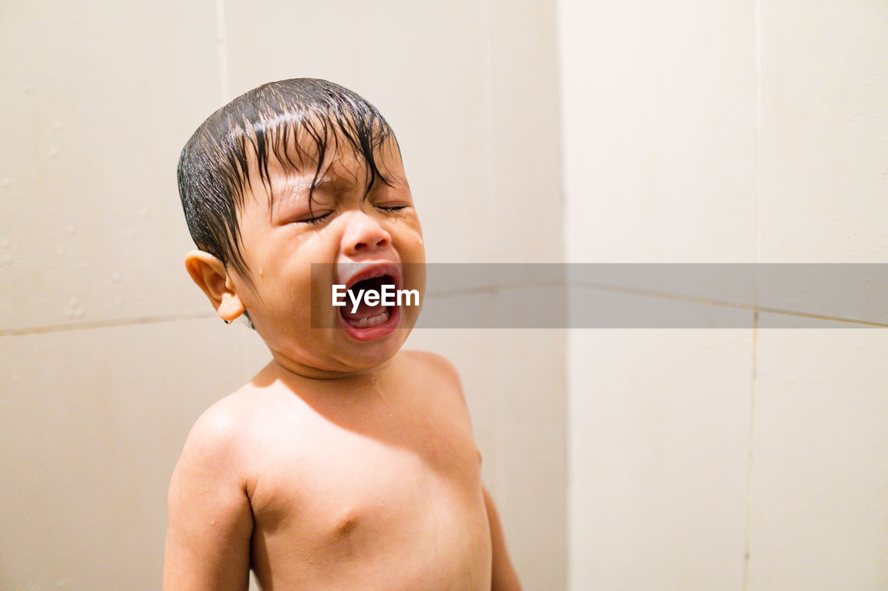 PORTRAIT OF SHIRTLESS BOY AGAINST WALL IN BATHROOM