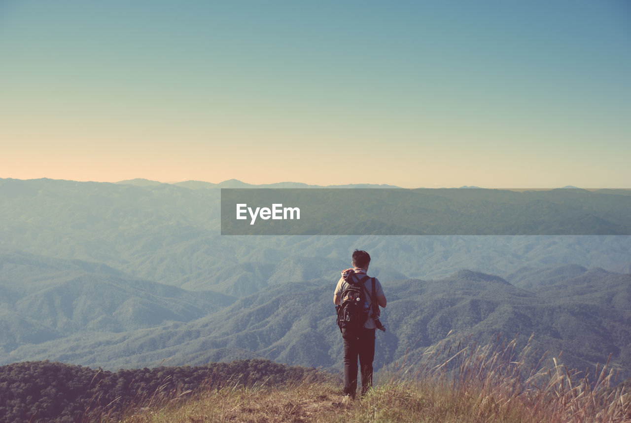 Rear view of young man standing on cliff against mountains