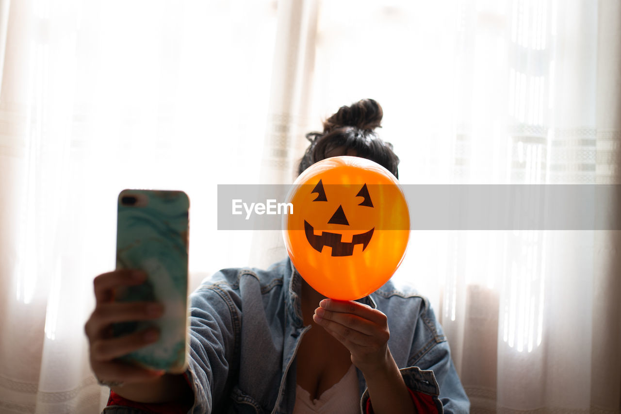 Close-up of woman holding halloween decoration against face and taking selfie at home